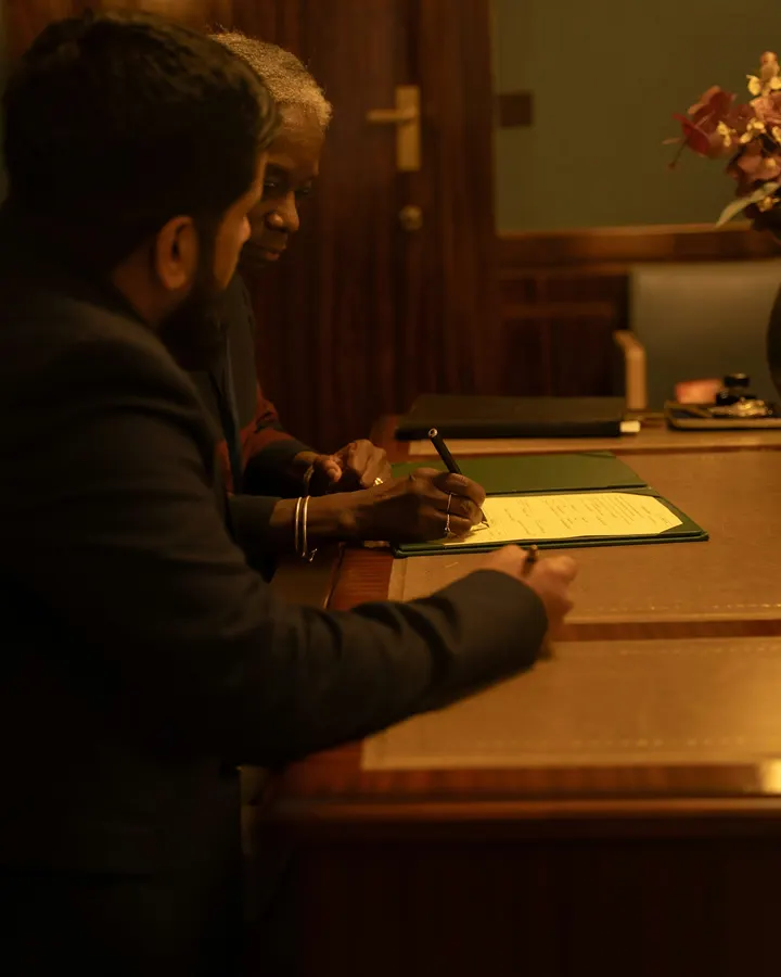 Two people signing papers across a desk in a wood-panelled chamber